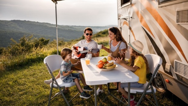 Happy family having fun while singing at picnic table by the camper trailer in nature. Man is playing a guitar.